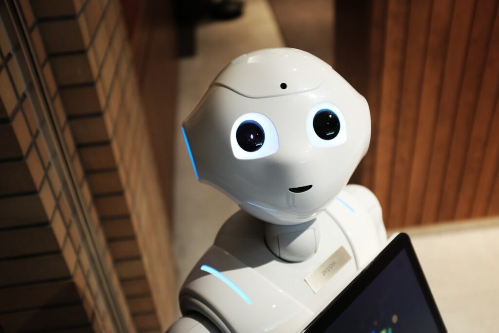 White humanoid Pepper robot with a friendly face and glowing blue accents stands in a hallway, screen partly visible in front.