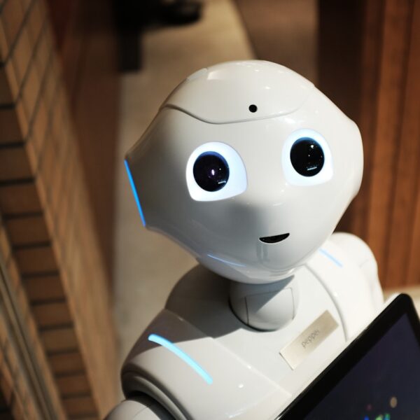White humanoid Pepper robot with a friendly face and glowing blue accents stands in a hallway, screen partly visible in front.