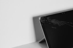Black-and-white photo of a laptop with a cracked screen propped up on a desk edge.